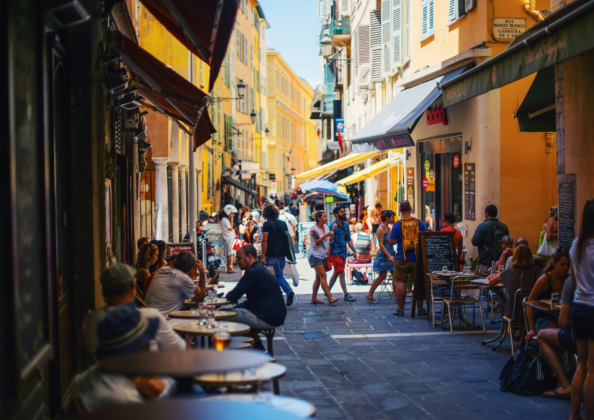Rue animée du Vieux-Nice, bordée de façades colorées et de cafés en terrasse. Les passants se croisent dans cette ruelle étroite et chaleureuse, typique de l’ambiance niçoise, entre commerces locaux, restaurants et vie de quartier ensoleillée.