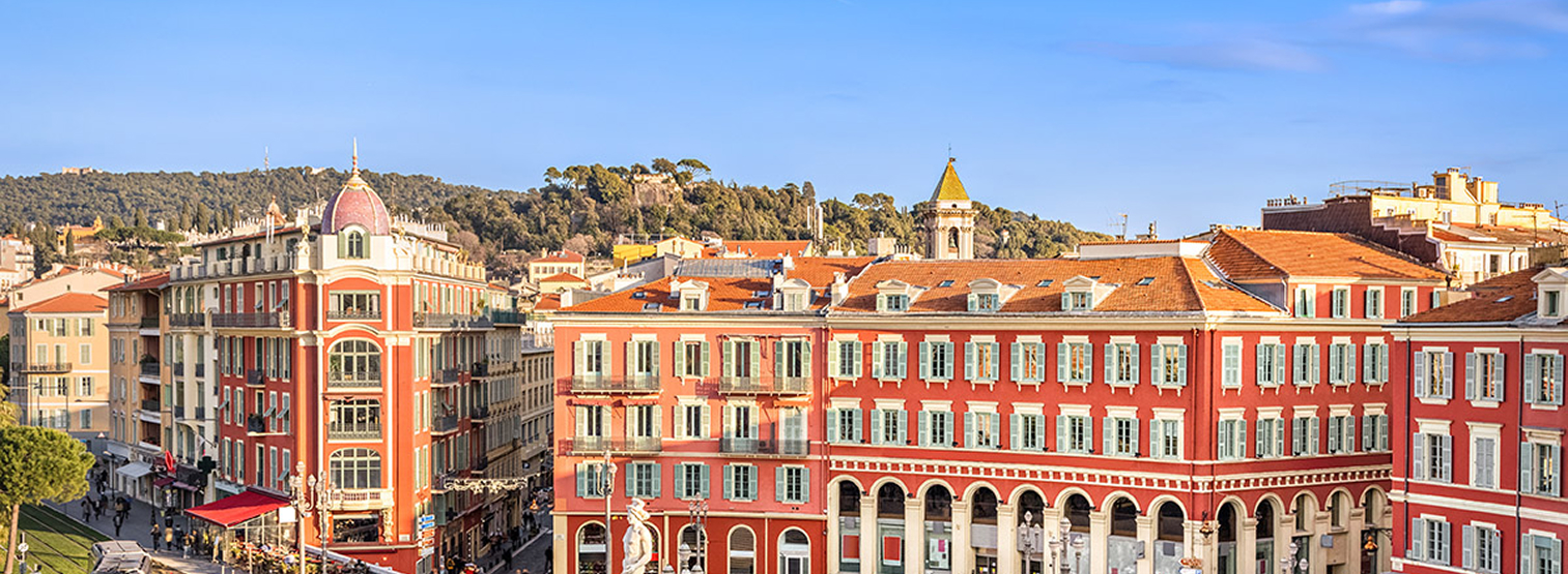 Vue panoramique d’un paysage urbain européen avec des bâtiments historiques colorés aux façades rouges et aux détails blancs, sur fond de collines verdoyantes et d’un ciel bleu clair. Un clocher et plusieurs toits en tuiles de terre cuite sont visibles.