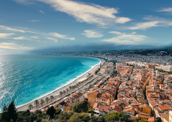 Vue aérienne spectaculaire du littoral niçois, où la Méditerranée d’un bleu intense longe la vieille ville aux toits orangés. La promenade bordée de palmiers s’étire le long de la plage, offrant un panorama emblématique entre mer, collines et architecture méditerranéenne.